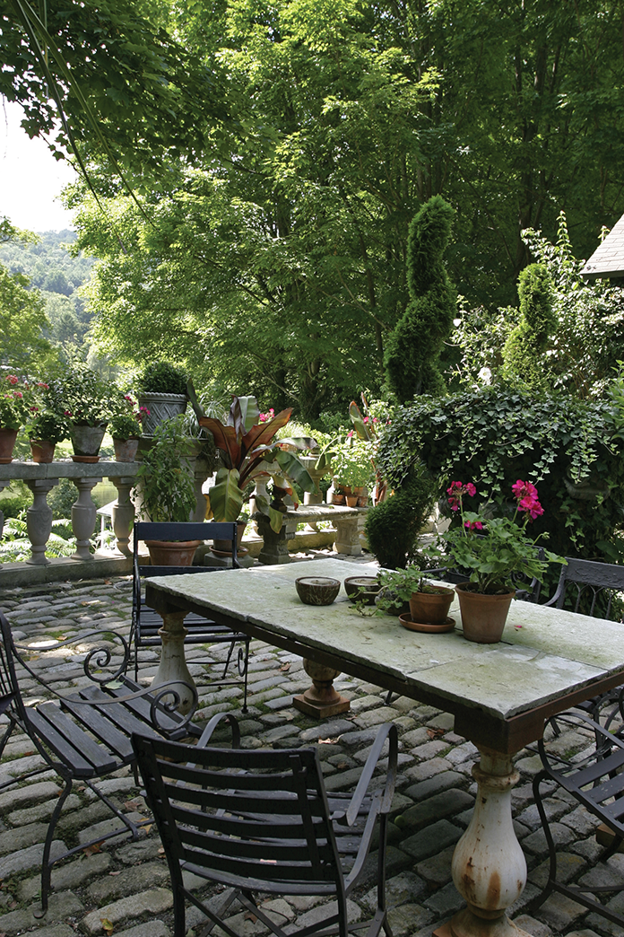 The back terrace includes a stone table made from antique flooring and French chairs. Potted plants surround the perimeter.